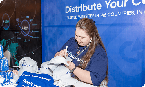 Membre de l'équipe Collaborator en chemise de marque signant des t-shirts promotionnels et des supports à un stand au SEO Mastery Summit, avec le message de distribution de contenu mondial de Collaborator visible en arrière-plan